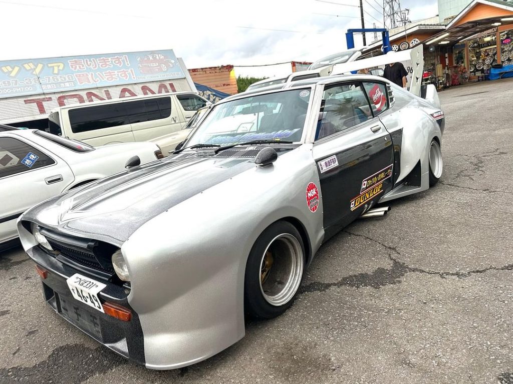 Modified retro Japanese car with wide body kit and race decals in a parking lot, store signage in Japanese background.