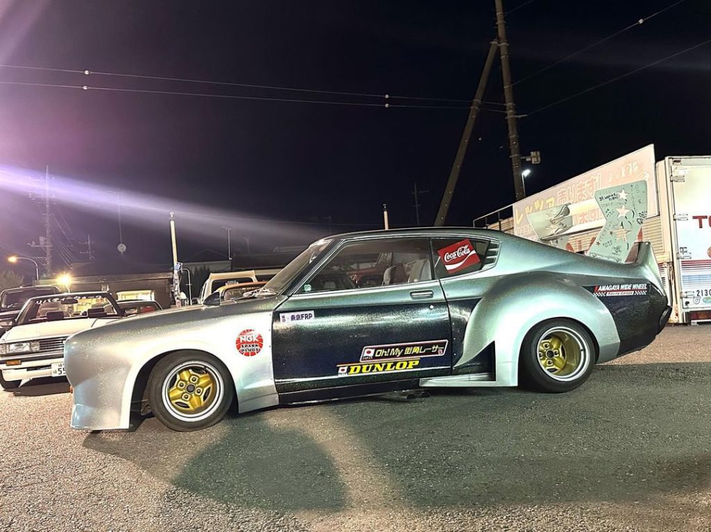 Vintage Japanese sports car with Dunlop logos, NGK sticker, and Coca-Cola decal at a night car meet.