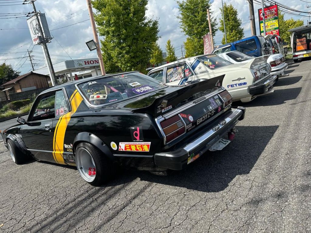 Classic Japanese cars lined up with detailed custom decals and modifications, parked near a Honda dealership.