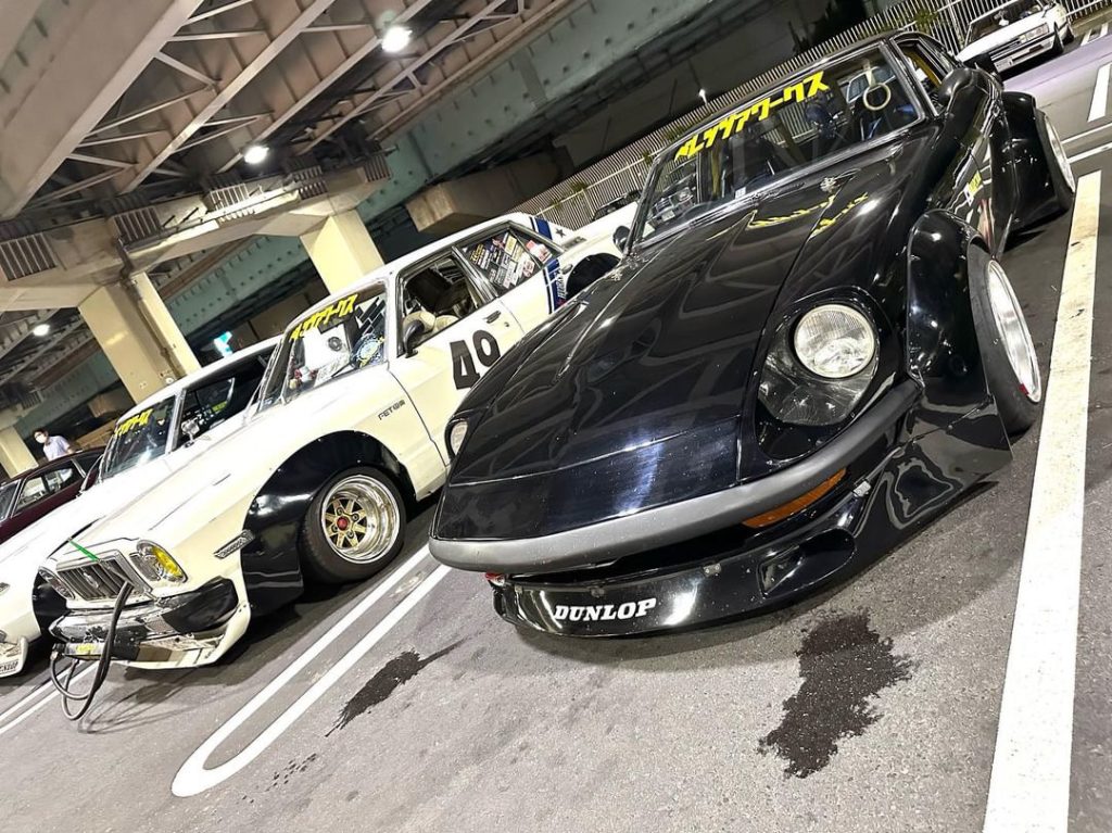 Classic Japanese tuner cars on display under an overpass. Prominent signage includes Dunlop and レディーカーズ (Lady Cars Club).