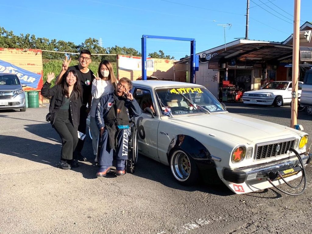Group posing with a vintage car at a garage. Japanese text on windshield, クラシックカー translates to Classic Car.