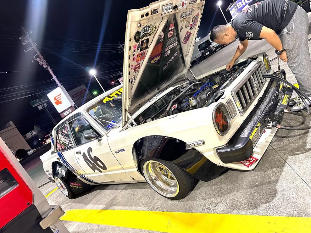 Mechanic working on vintage race car number 46 at a gas station at night in Japan with hood open, showcasing engine.