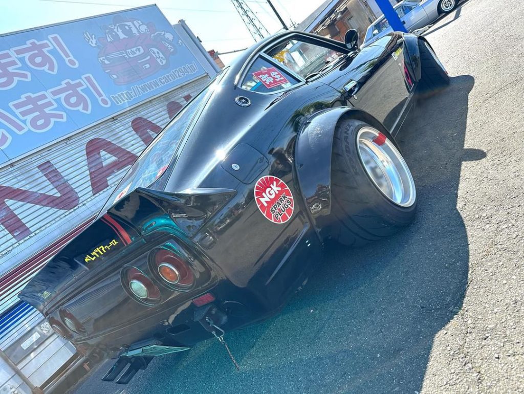 Black custom car with NGK sticker, modified body kit, and wide tires outside a garage with Japanese signage in the background.