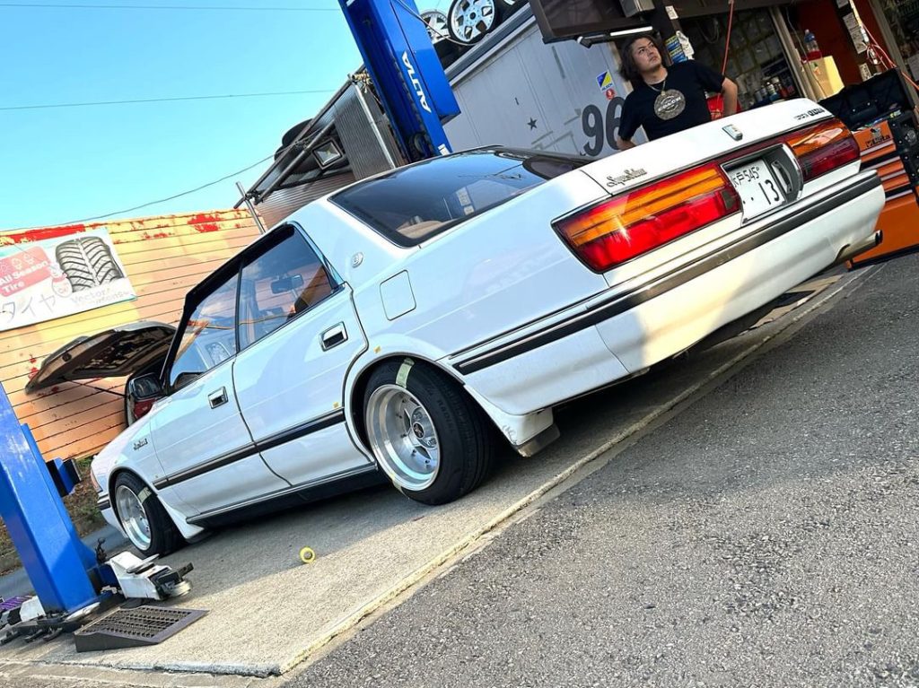 A white vintage Toyota Crown car on a lift at an auto repair shop. Sign reads All Seasons Tires in Japanese.