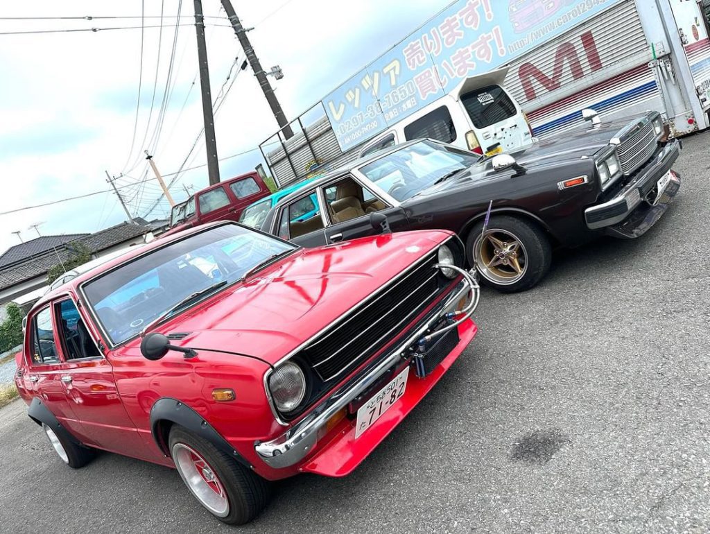 Two classic Japanese cars parked outside in a vintage car show setting, one red and one black, showcasing retro style.