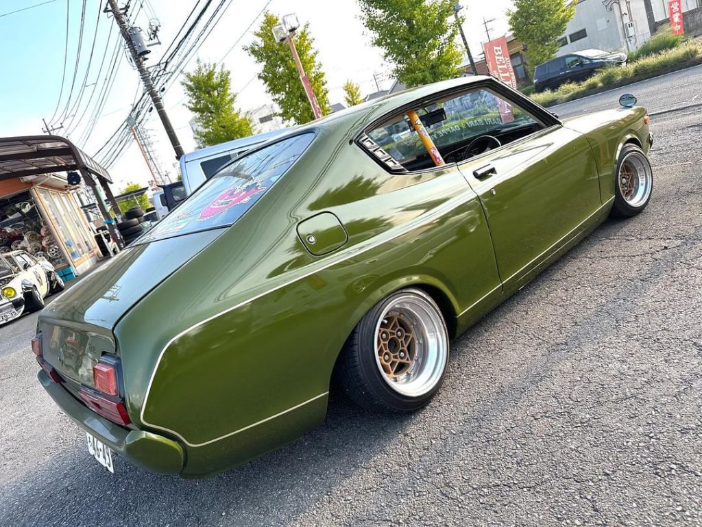 Green classic car with custom wheels parked on street near car garage in Japan. Clear sky and trees in the background.