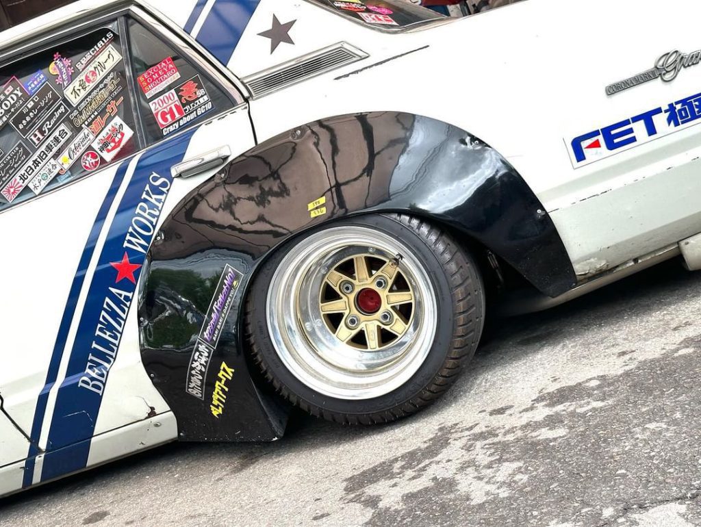 Side view of a modified retro Japanese car with decals, showcasing large custom wheels and vibrant graphics.