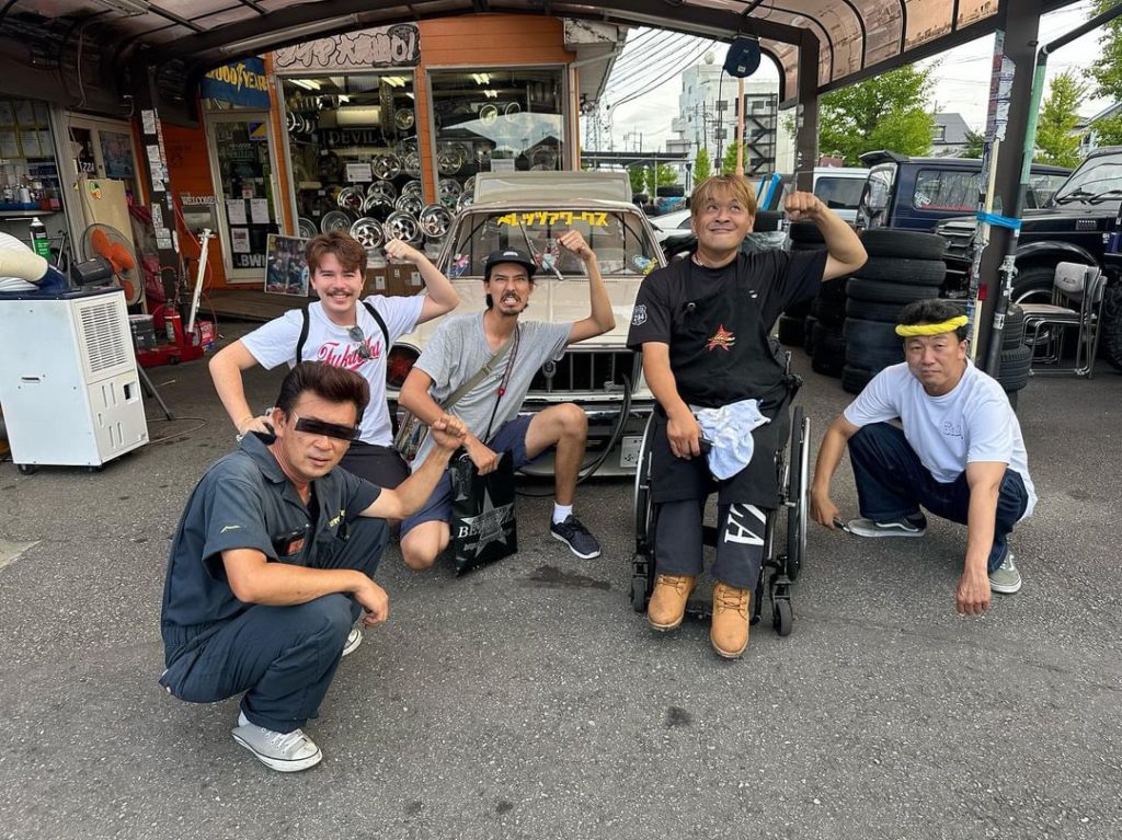A group of men posing flexibly in front of a car repair shop with Japanese signage and a display of car rims.