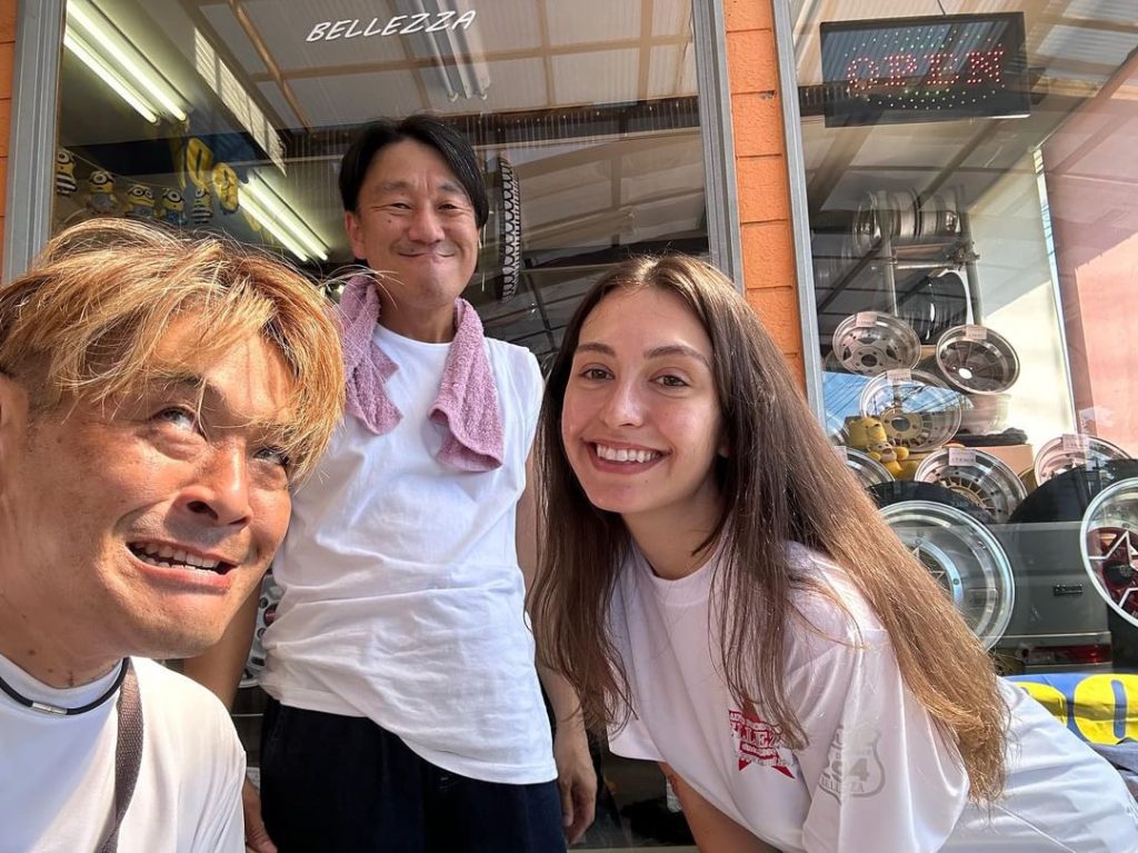 Three people posing and smiling in front of a shop with custom car rims and Bellezza and Open signs.