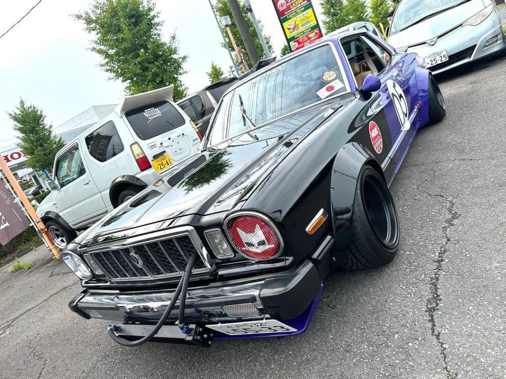 Modified classic Japanese car with sticker decals and black paint in a parked car meet-up.