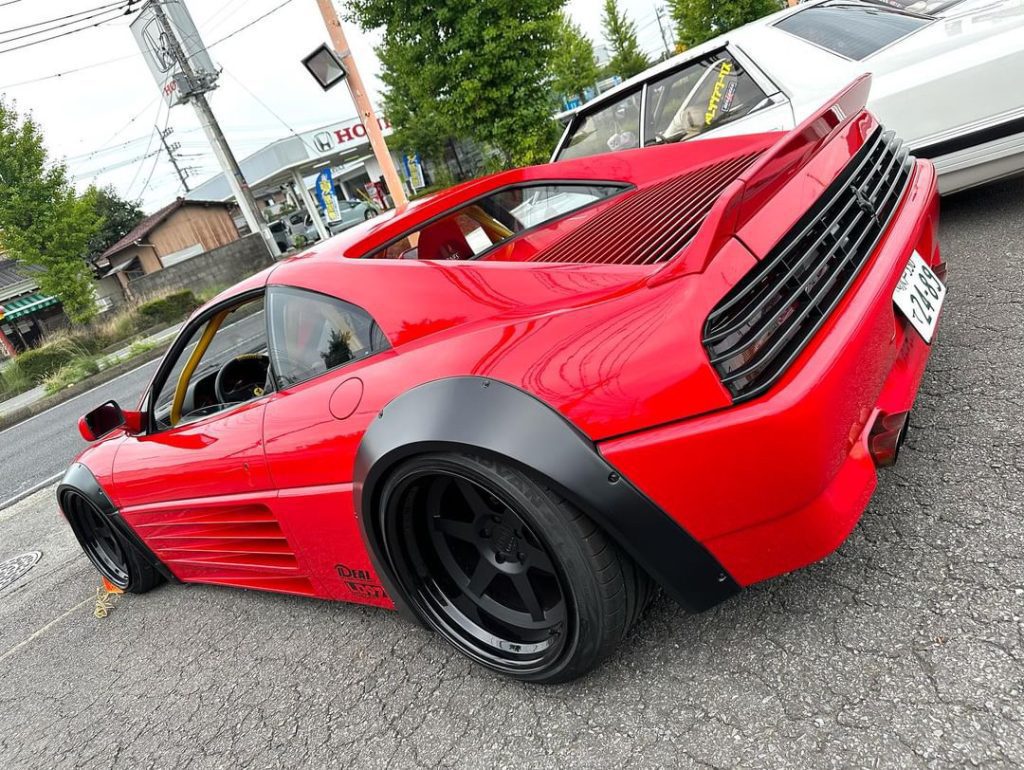 Red sports car with custom black wheels and rear wing parked on a street beside a white car near Honda dealership in Japan.