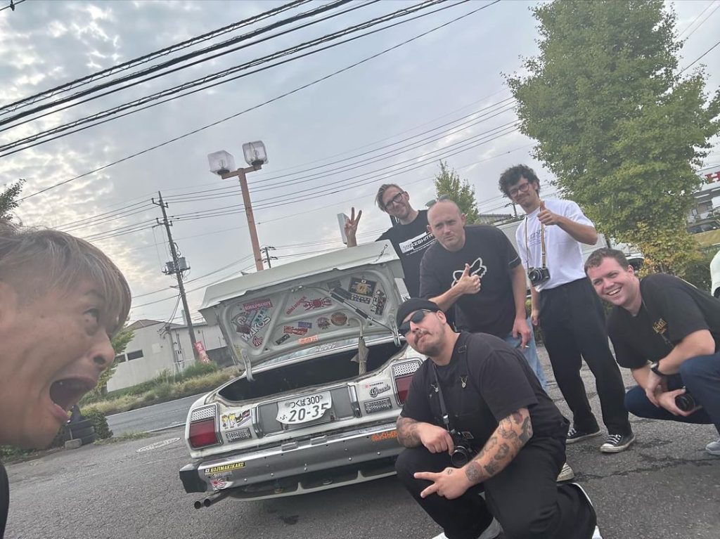 Group of friends posing by a retro car with license plate つくば300 20-35 in an urban area, one person makes a surprised face.