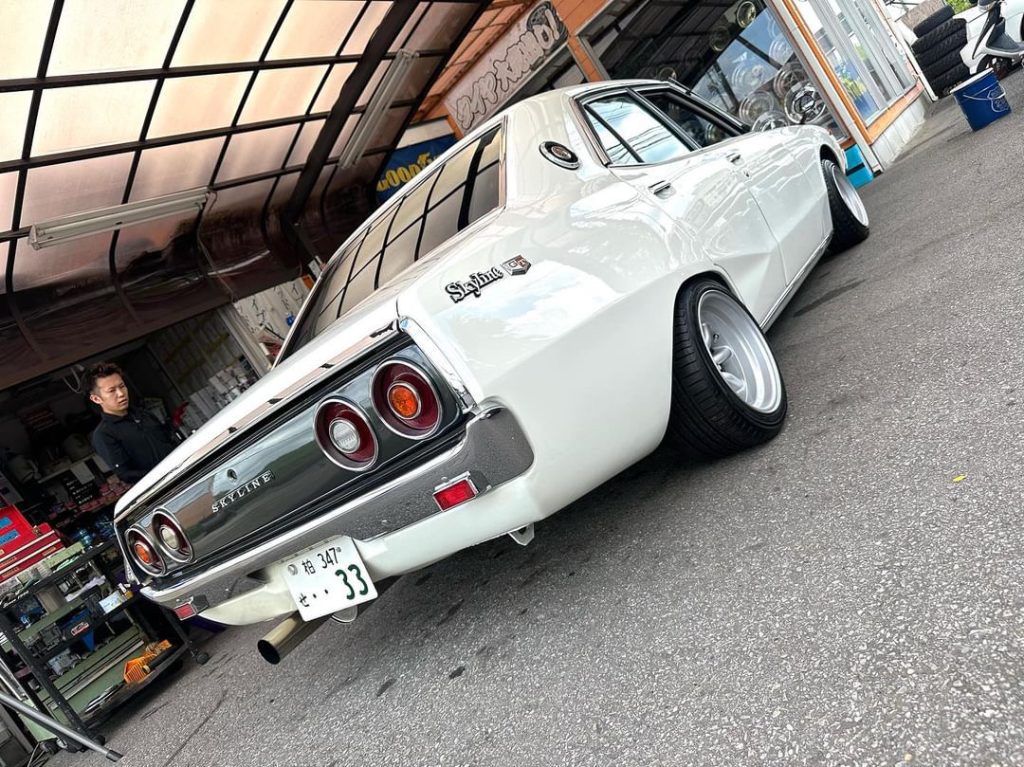 Classic white Nissan Skyline GT parked in a garage. Japanese license plate and Skyline emblem visible.
