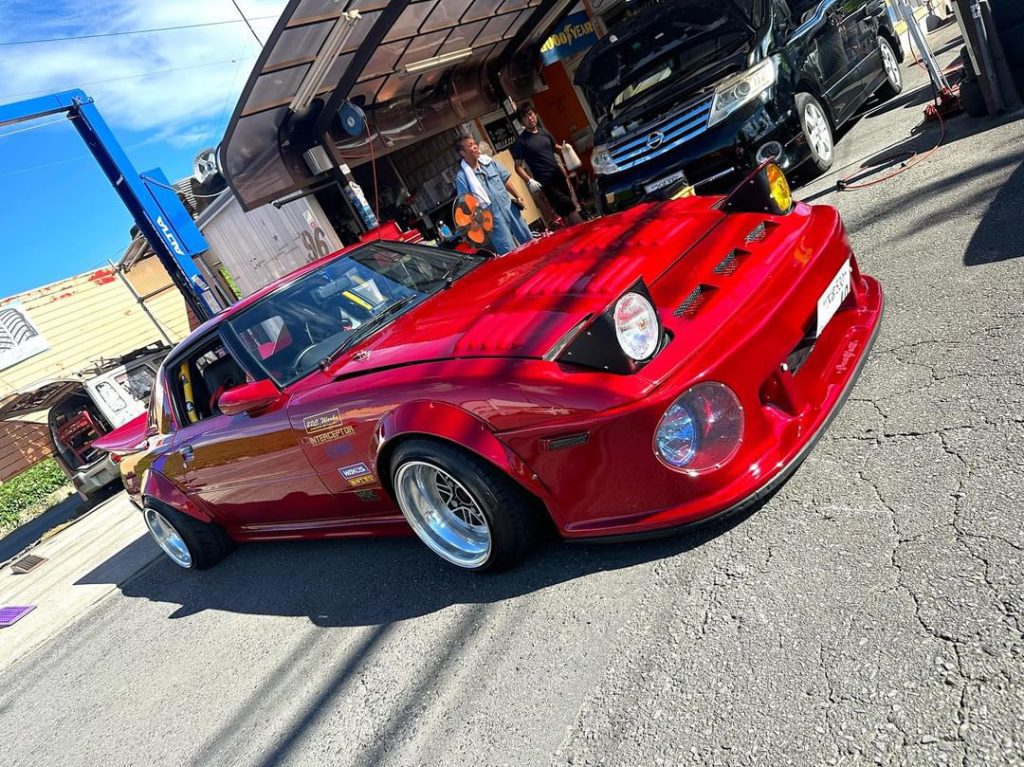 Red custom car at Japanese auto shop with mechanics, featuring detailed body modifications and polished rims.