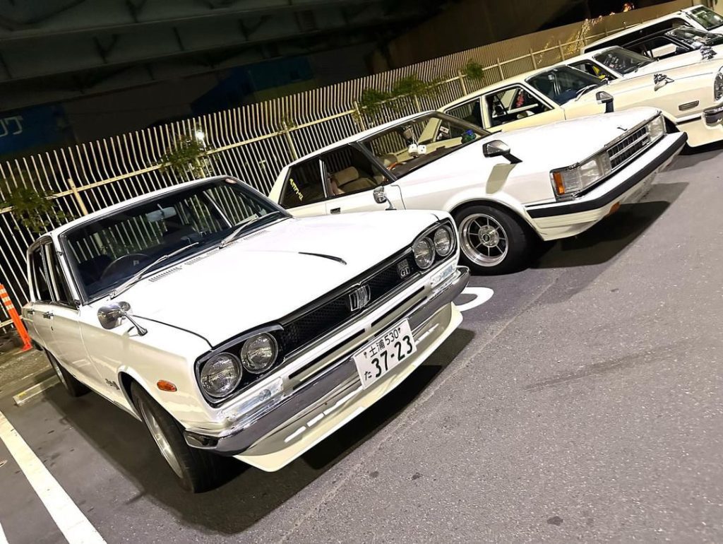 Vintage Japanese cars parked in a row at night. License plate 37-23 visible on the front car. Classic car meet.