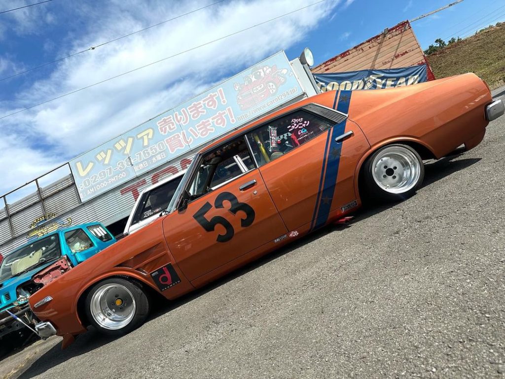 Vintage orange race car with number 53, blue racing stripes, parked outside a business with Japanese signage and a blue truck.