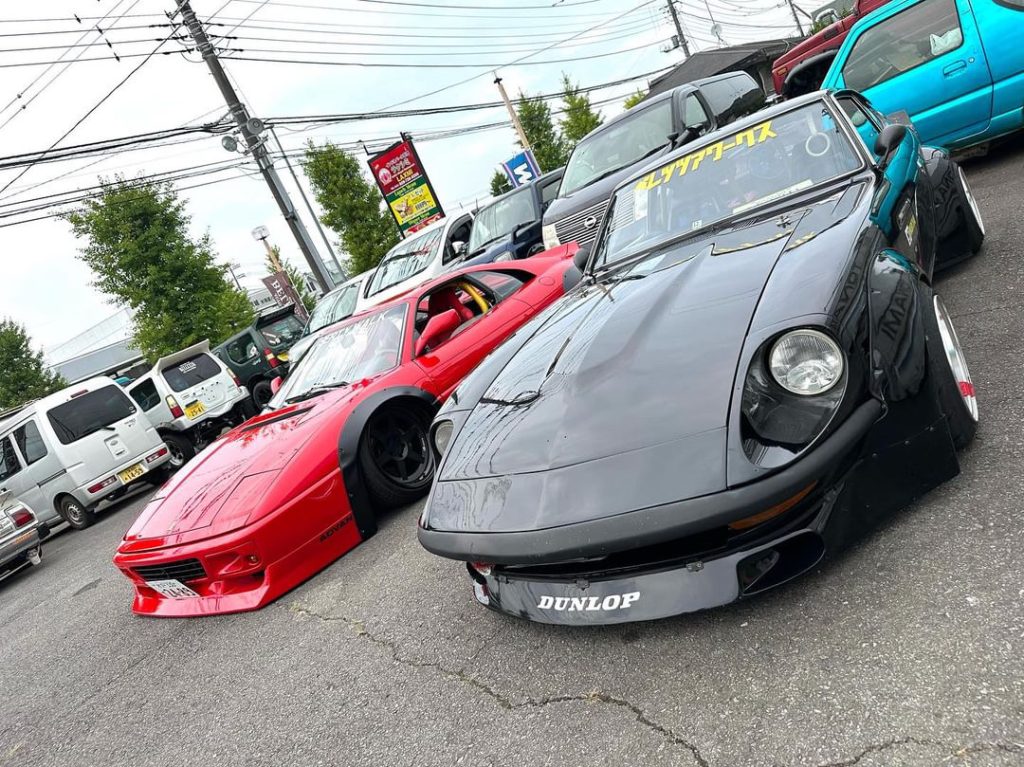 Two modified sports cars, a red Mazda RX-7 and a black Nissan Fairlady Z, parked in a lot, with Japanese signage.