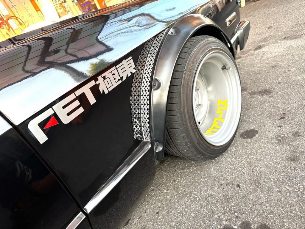 Close-up of a black car with the decal FET 極東 (Far East) and custom alloy wheels on a city street.