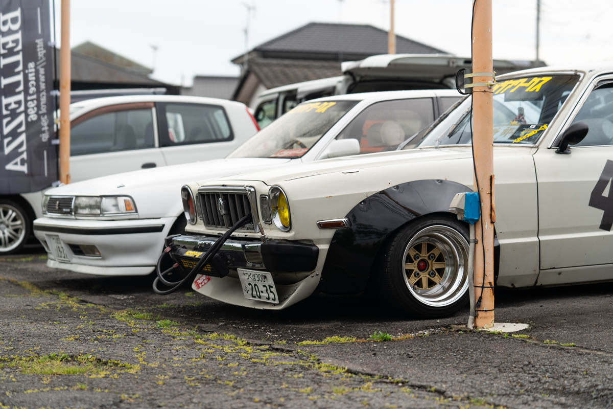 Vintage modified Japanese car with wide fender flares, parked at an event. Other cars and a banner in the background.
