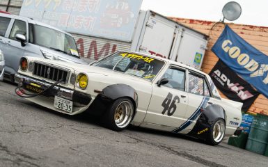 Classic Japanese car with Yokohama Advan livery, parked by a garage; racing number 46; yellow headlights; retro-mod style.