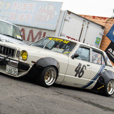 Classic Japanese car with Yokohama Advan livery, parked by a garage; racing number 46; yellow headlights; retro-mod style.