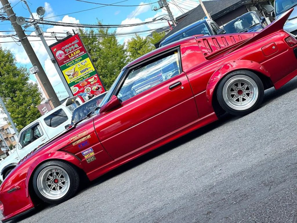 Red modified sports car with custom decals parked outside a restaurant in Japan. Japanese menu sign visible in background.