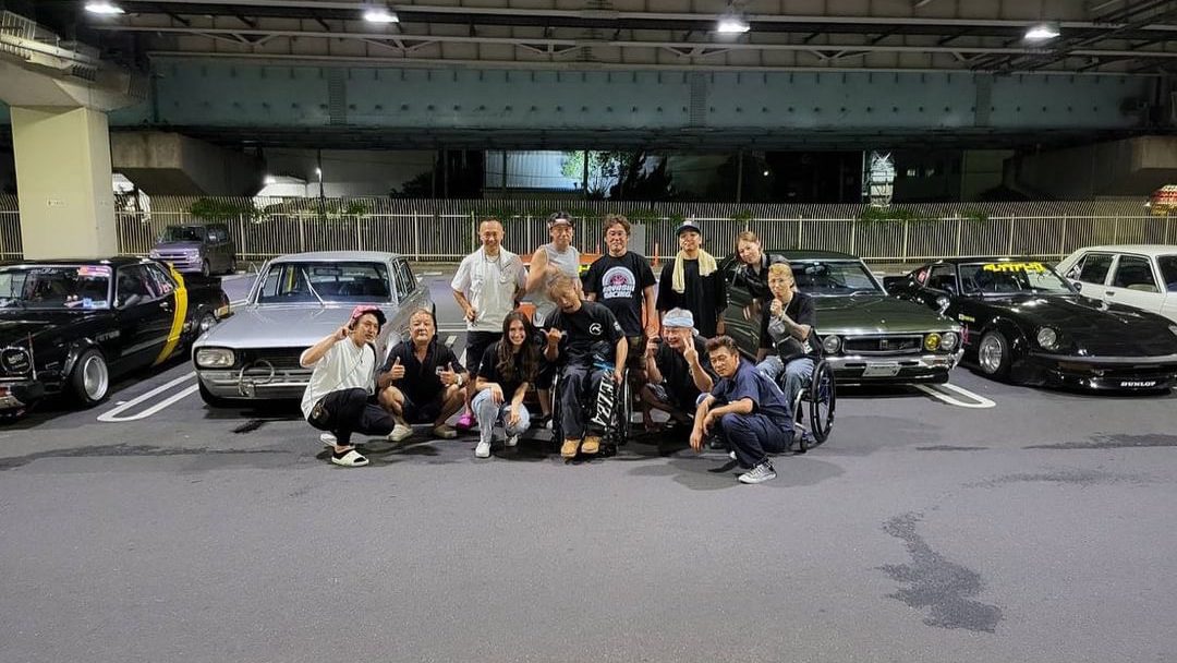 A group of people posing with vintage cars in a parking lot under a bridge at night in Japan, showing camaraderie.