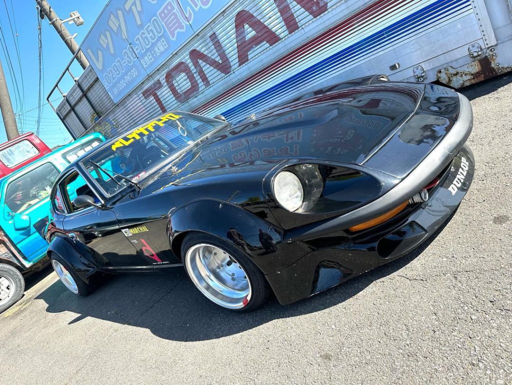 Classic black sports car with aftermarket mods and yellow windshield banner at Tonan garage in Japan, next to blue van.