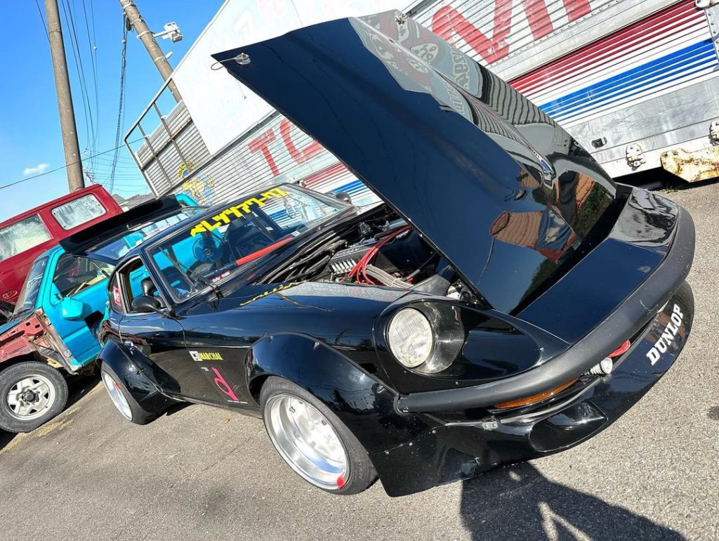 Modified black sports car with hood up, Dunlop sticker, parked next to blue vehicle, sunny day, power lines in background.