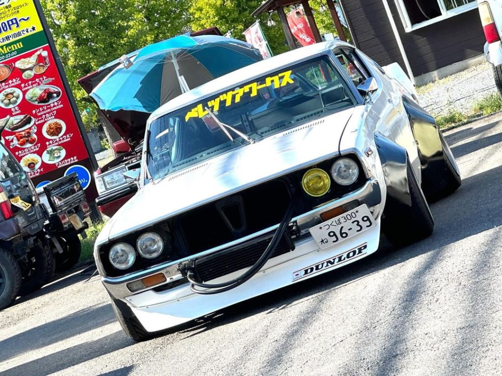 Modified classic Japanese car with a Dunlop bumper sticker and a Japanese license plate in front of a food menu sign.