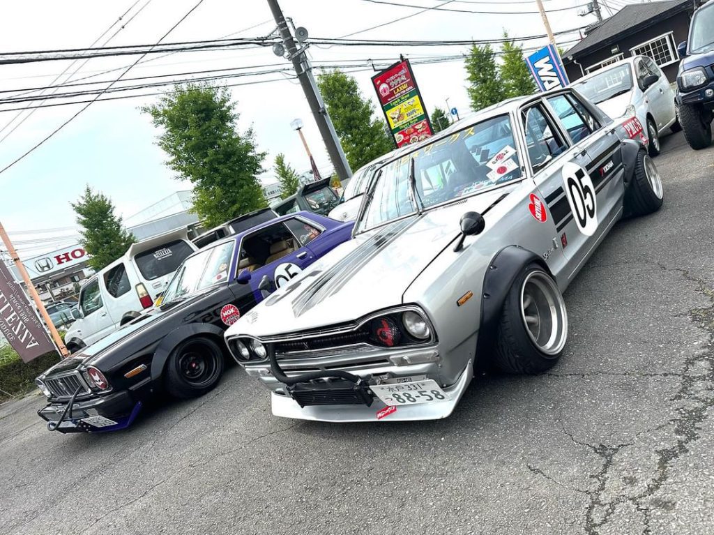 Two modified vintage Japanese cars in a parking lot; a silver car with the number 05 and a black car with a roll cage.
