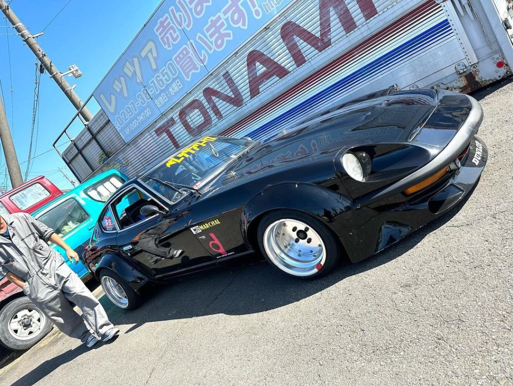 Classic black sports car parked near Tonami sign in Japanese, with a man in gray overalls and a turquoise vehicle in the background.