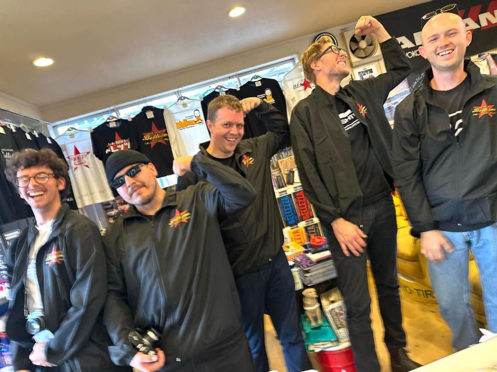 Group of five men in black jackets posing and smiling inside a store with merchandise in the background.