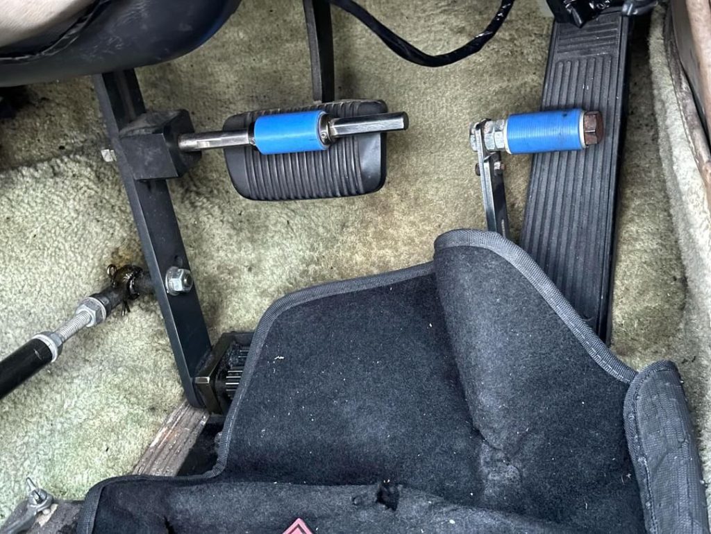 Close-up of vehicle foot pedals with blue modifications and carpet mat, showcasing the accelerator and brake pedals.