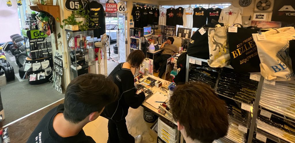 People shopping in a Japanese store filled with car accessories, apparel, and bags, with shirts displayed on the walls.