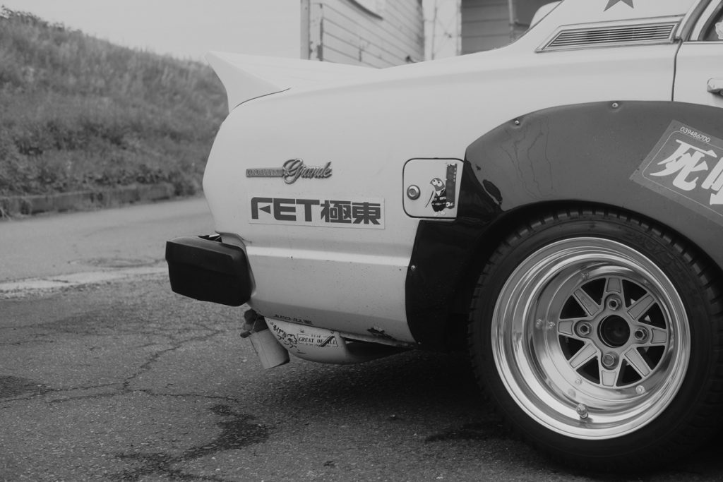 Close-up of a classic car's rear side, featuring detailed alloy wheels and retro Japanese decals on the body.