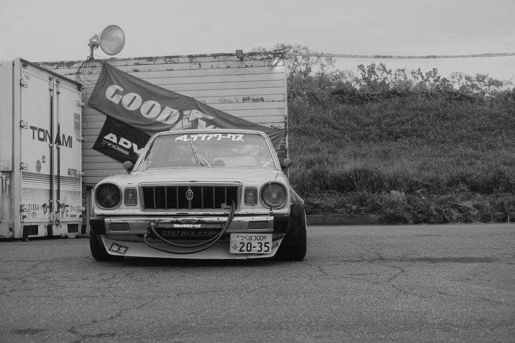 Vintage Japanese car parked in front of Good and ADVAN Yokohama banners with a lowered suspension and license plate.