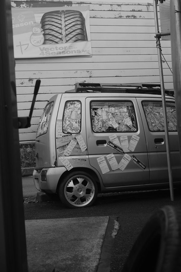 A parked car covered with advertisements and notices, below a poster promoting all-season tires in Japanese.