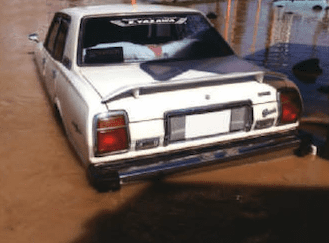 White car partially submerged in floodwaters, rear view with Japanese sticker on window.
