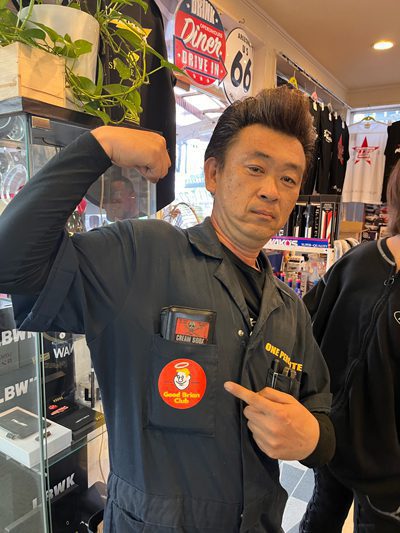 Man in a shop displaying a Good Ramen Club sticker and a Cuban Snook phone case from his pocket, with Route 66 decor behind.