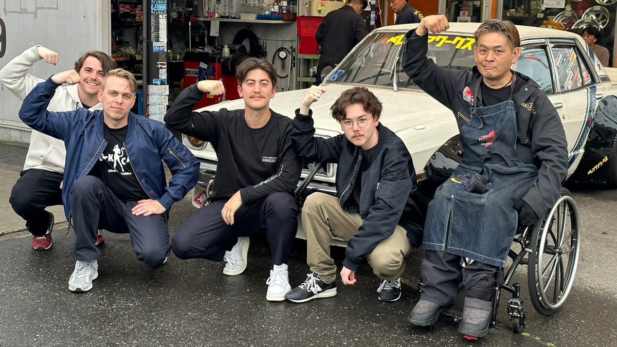 Group of five men posing and flexing biceps in front of a customized car, Japanese text on windshield reads メタルワークス (Metal Works).