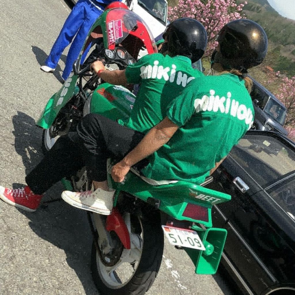 Two riders on a green motorcycle with matching green shirts that say makino, parked near blooming cherry blossoms.