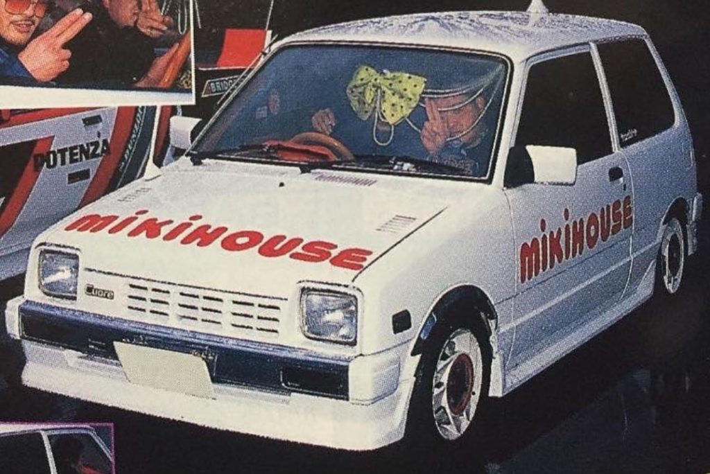White car with Mikihouse branding, custom rims, bow on windshield, and people inside at an auto show.