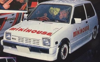 White car with Mikihouse branding, custom rims, bow on windshield, and people inside at an auto show.