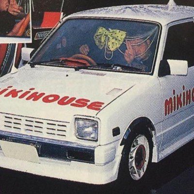 White car with Mikihouse branding, custom rims, bow on windshield, and people inside at an auto show.