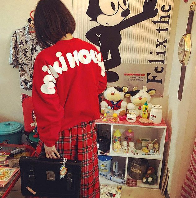 Young person in red MIKI HOUSE sweatshirt and plaid skirt, standing by vintage Felix the Cat posters and plush toys.