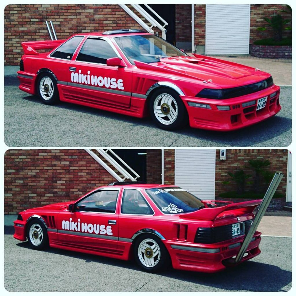Red modified sports car with Miki House branding and large exhaust pipe, parked outside a brick building.