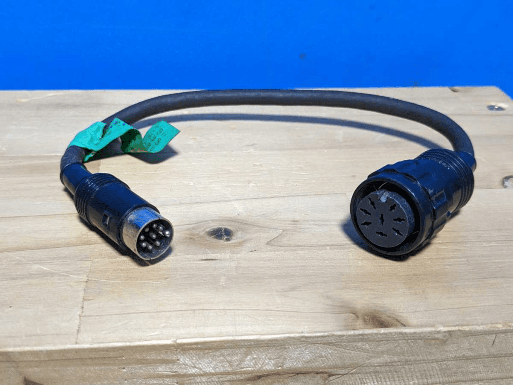 Close-up of a black 6-pin DIN cable connector on wooden surface with blue background, green tag with Japanese writing.
