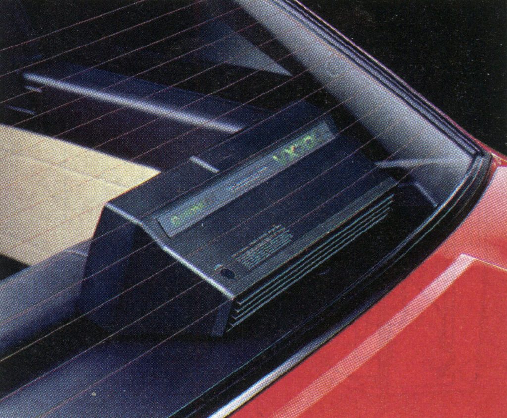 Alt text: Vintage Japanese rear car audio system with illuminated display in the back window of a red vehicle.