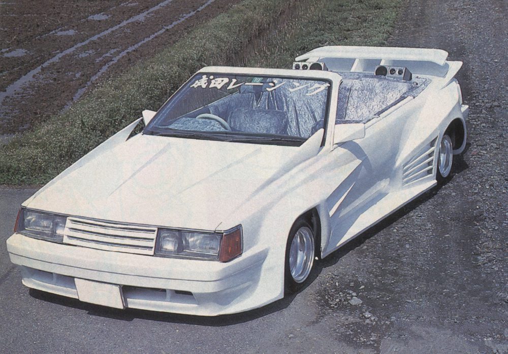 A modified white convertible car with custom body kit and the Japanese text 成田レンジャー on windshield, parked on a rural road.
